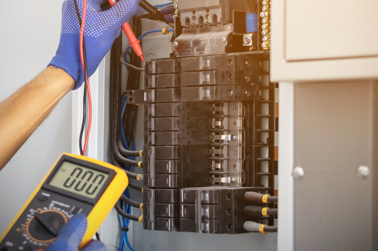 Électricien utilisant un multimètre pour tester un disjoncteur dans un tableau électrique ; gants bleus, multimètre jaune.