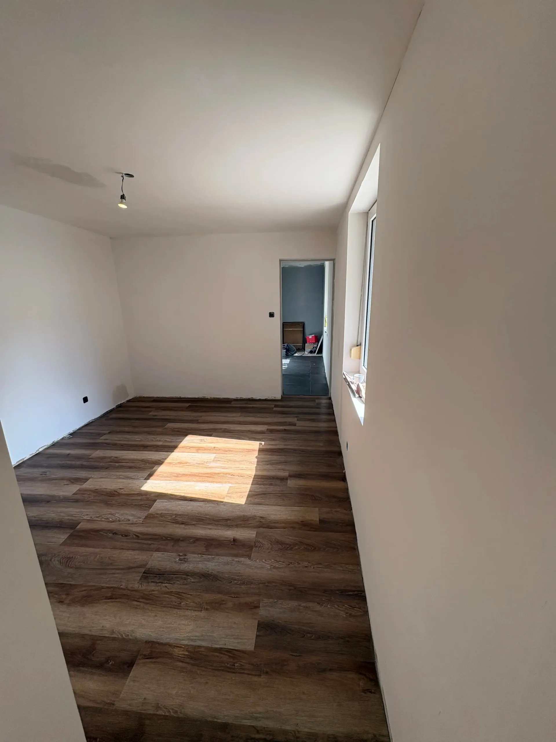 Pièce vide avec sol imitation bois, murs blancs et une fenêtre. Une porte ouverte donne sur une autre pièce.