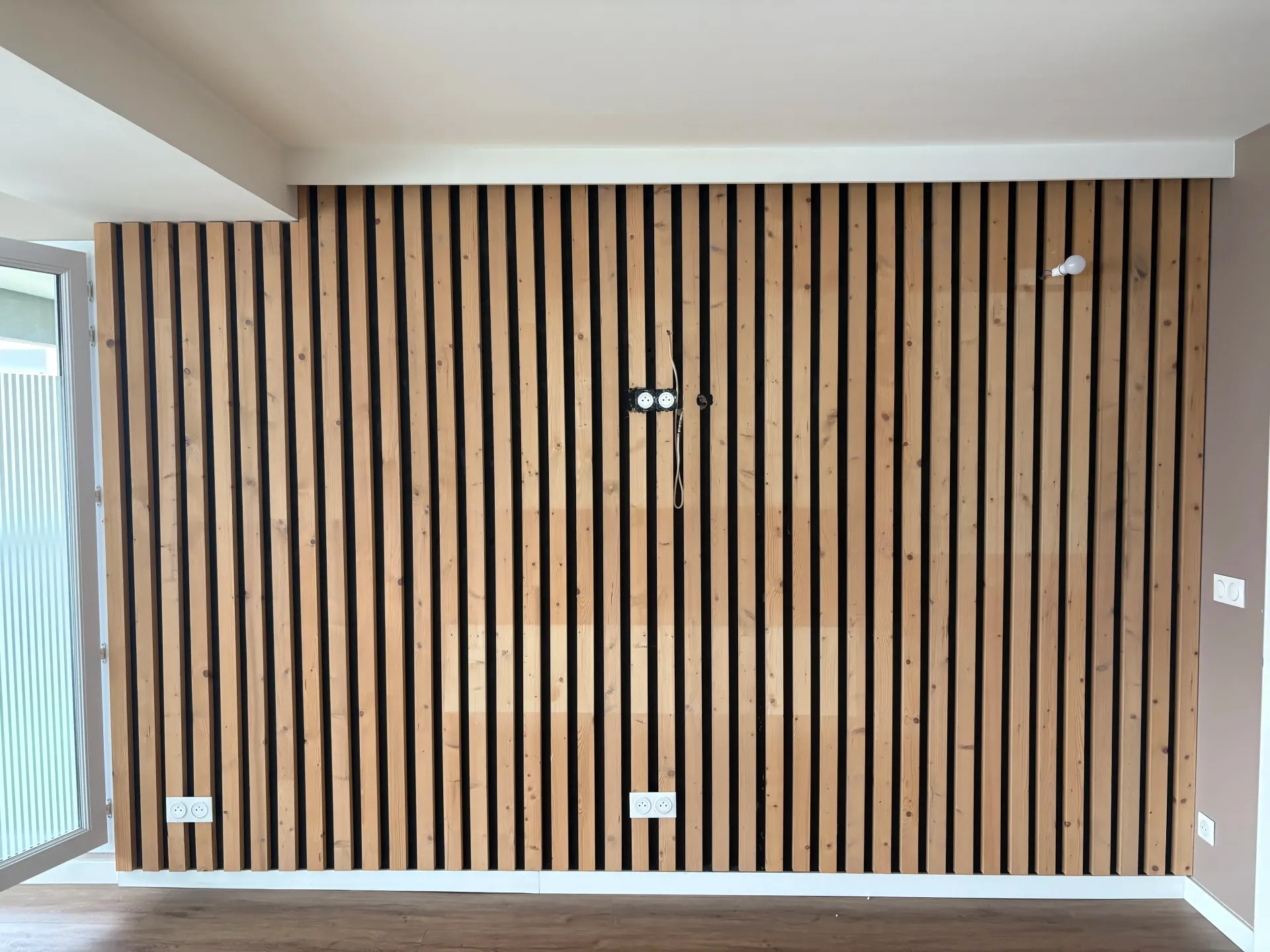 Cloison en lattes de bois avec prises électriques, moulures blanches et plafond blanc.