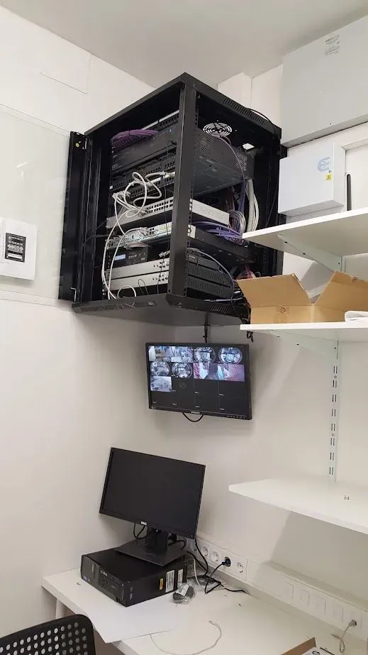 Una computadora está sobre un escritorio en una habitación al lado de un televisor montado en la pared.