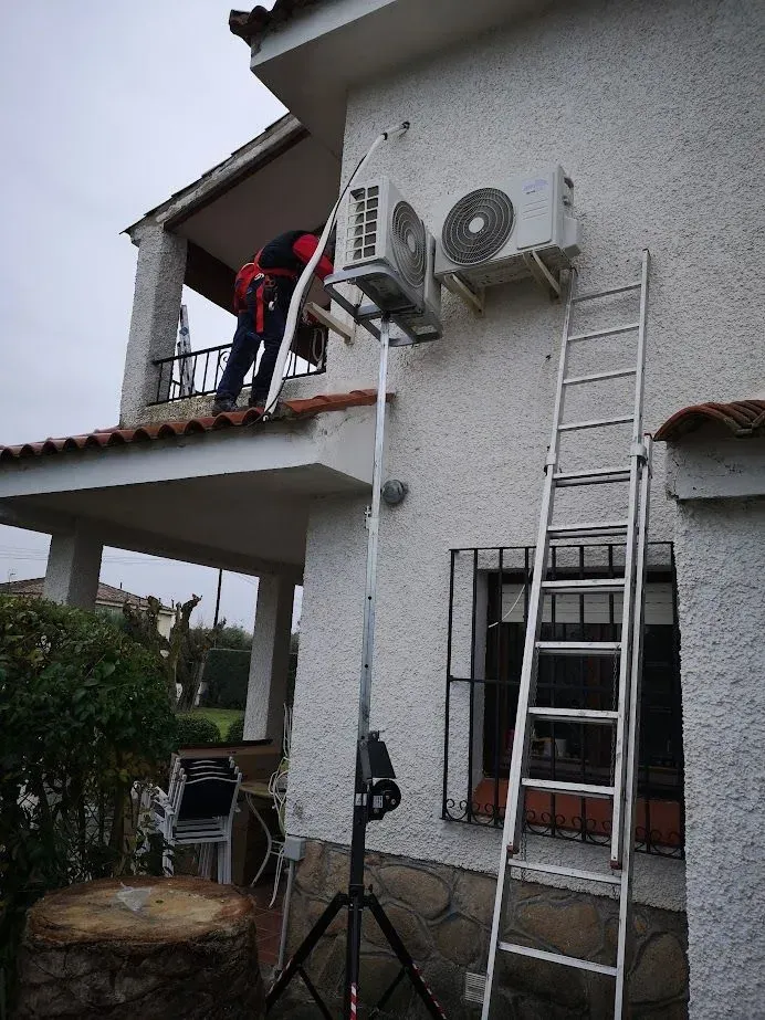 Un hombre en una escalera está trabajando en un acondicionador de aire al costado de una casa.