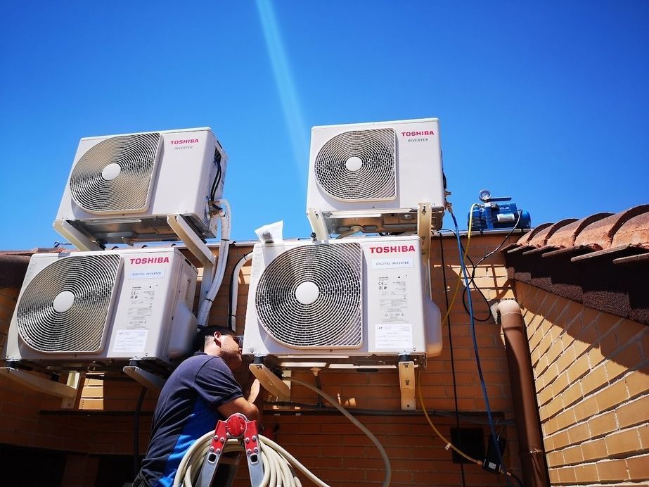 Un hombre está de pie en una escalera trabajando en acondicionadores de aire en el costado de un edificio.