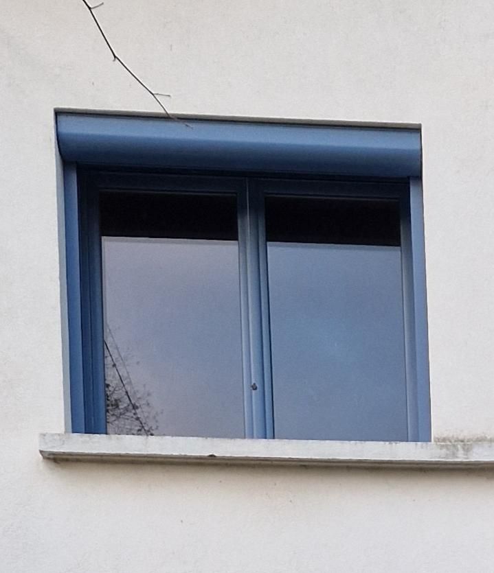 Fenêtre à cadre bleu avec volet enroulé, encastrée dans un mur blanc.