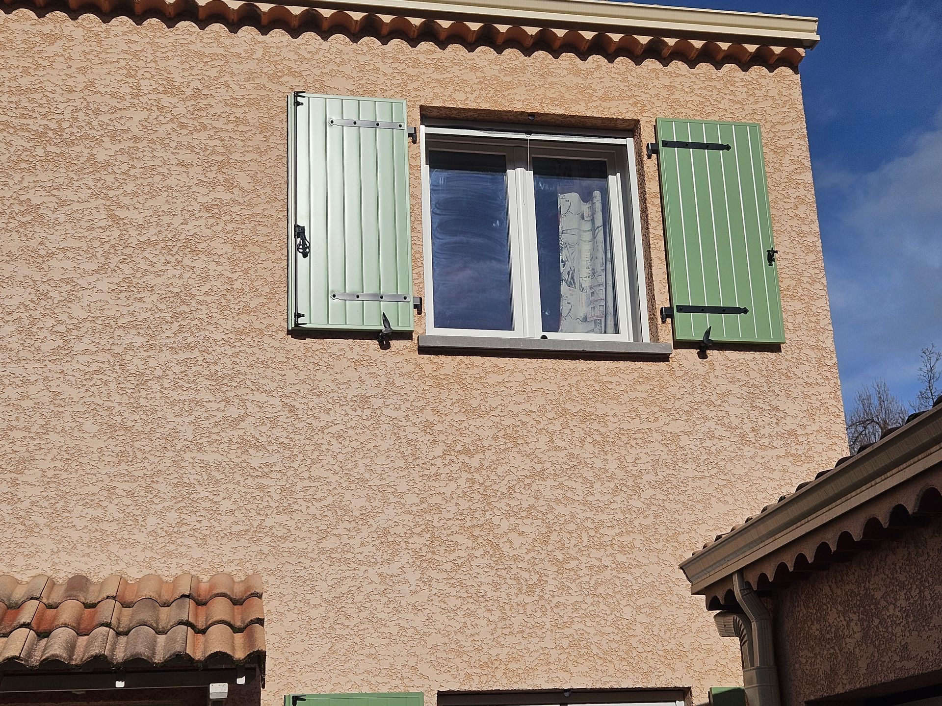 Fenêtre avec des volets verts sur un bâtiment en stuc ; le toit en dessous est en tuiles rouges.