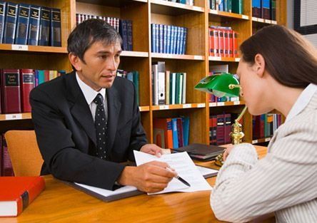 Dos personas en una biblioteca discutiendo artículos en una mesa de madera, con estanterías de libros al fondo.