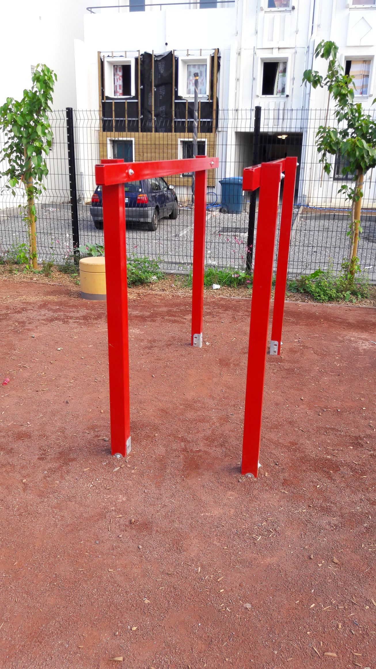 Barres de fitness rouges en extérieur, installées sur une zone de gravier dans un parc, avec des immeubles d'appartements en arrière-plan.