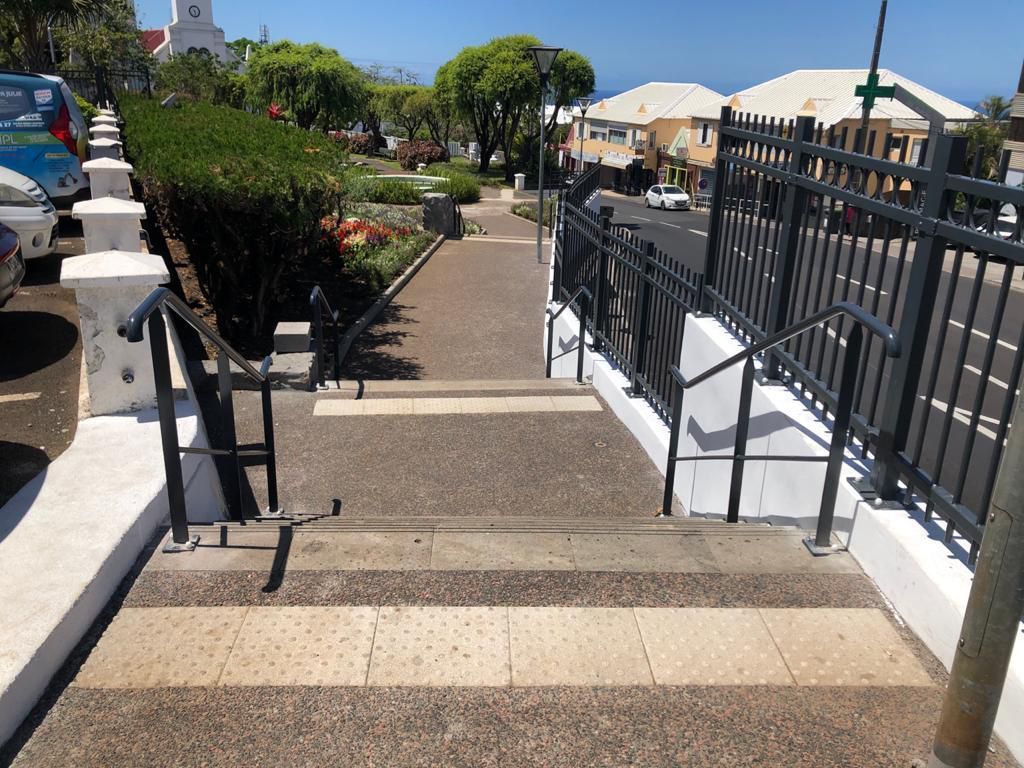 Rampe d'accès avec mains courantes menant à une terrasse extérieure ensoleillée et à une allée près d'une clôture