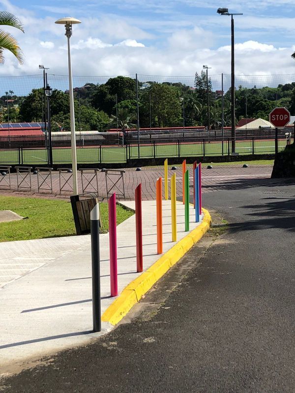 Des bornes colorées bordent un trottoir, à côté d'un chemin asphalté et d'un terrain de sport clôturé, sous un ciel nuageux.