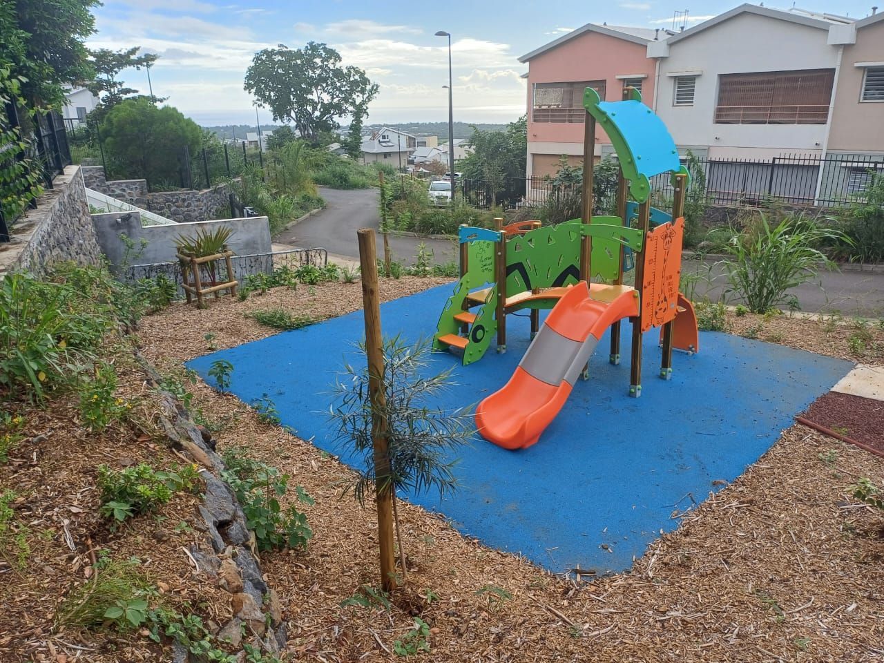 Petite aire de jeux avec toboggan coloré et équipements d'escalade sur tapis de sécurité bleu dans une cour résidentielle