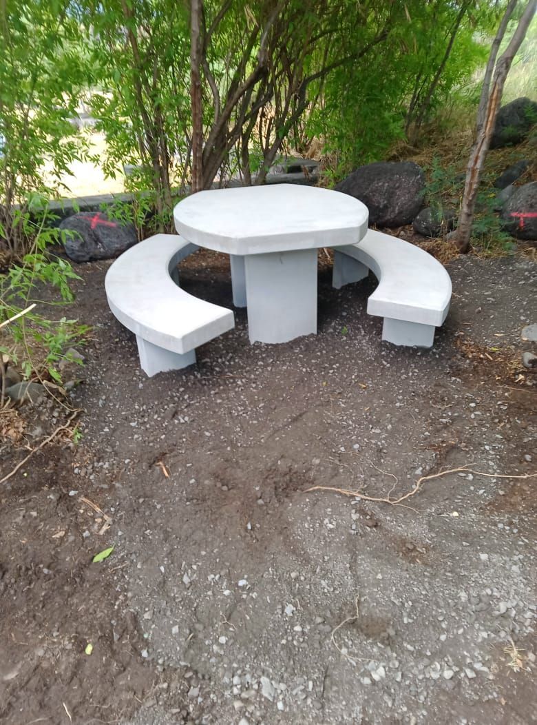 Table de pique-nique extérieure en béton blanc avec bancs incurvés sous les arbres, sur un sol en terre battue