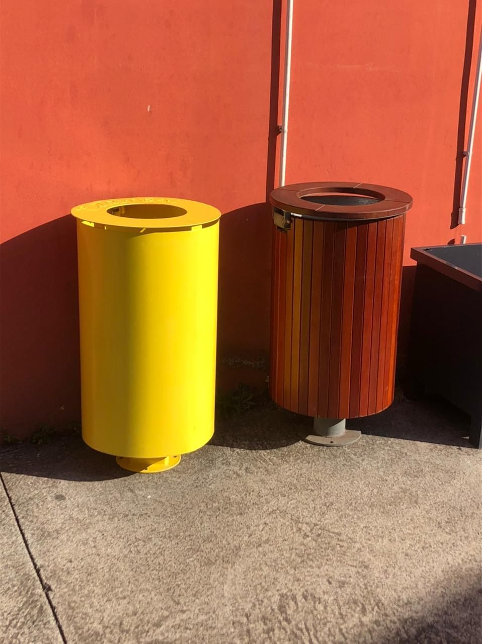 Des poubelles jaunes et imitation bois sont placées à côté d'un mur rouge sur un trottoir en béton.