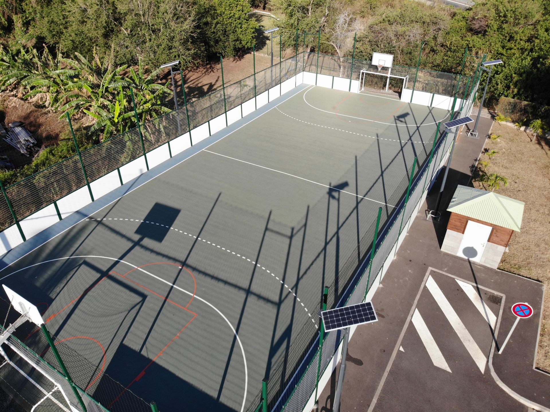 Terrain de sport extérieur vide, entouré d'une clôture, avec un banc, des arbres et un petit bâtiment à proximité.