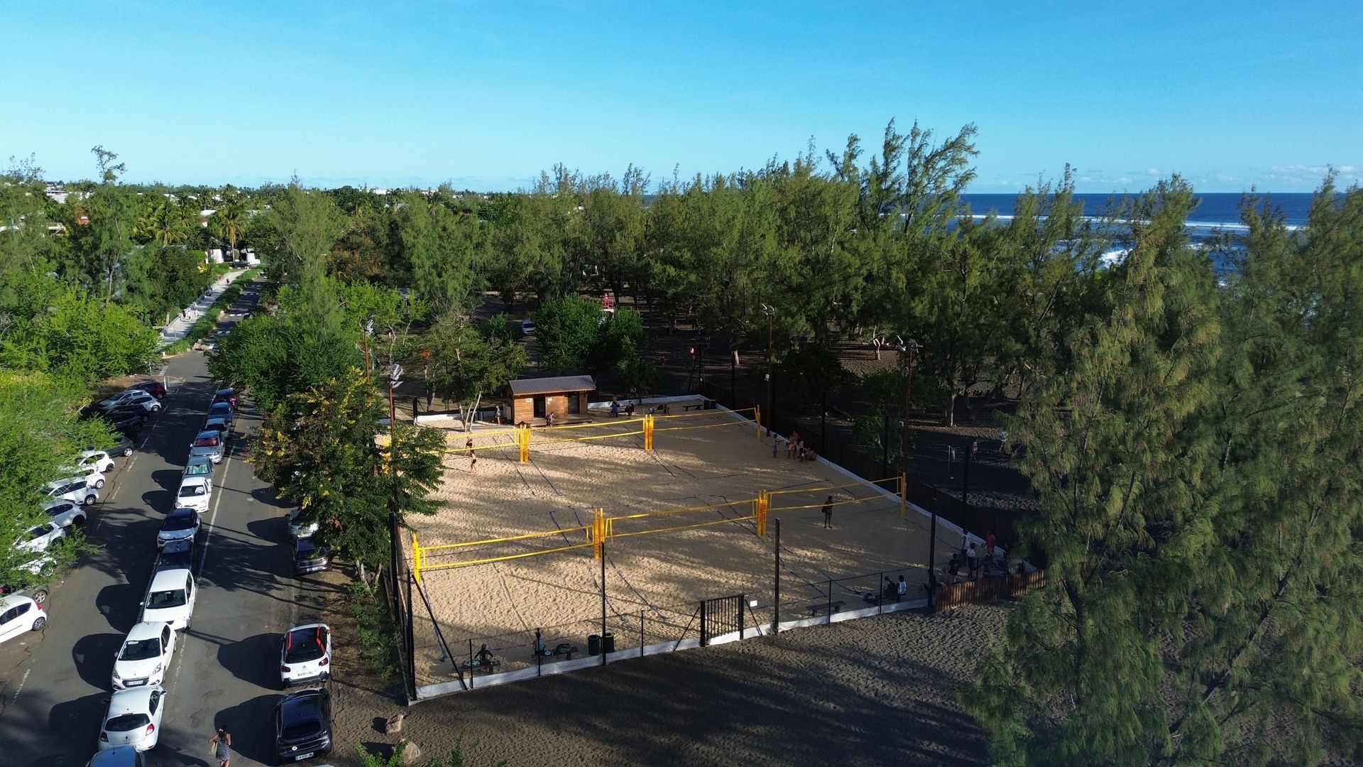 Terrain de beach volley extérieur clôturé en construction, entouré d'arbres, avec la mer visible en arrière-plan.