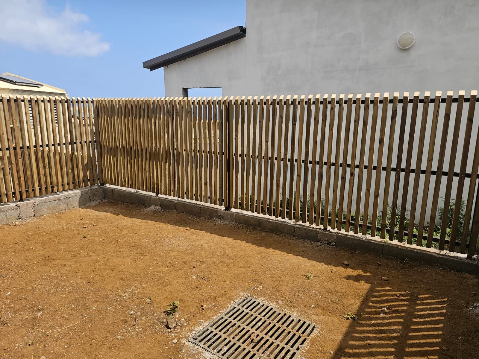 Petit jardin en terre battue clôturé avec un mur en béton, une clôture en lattes de bois et une grille d'évacuation, baigné de soleil.