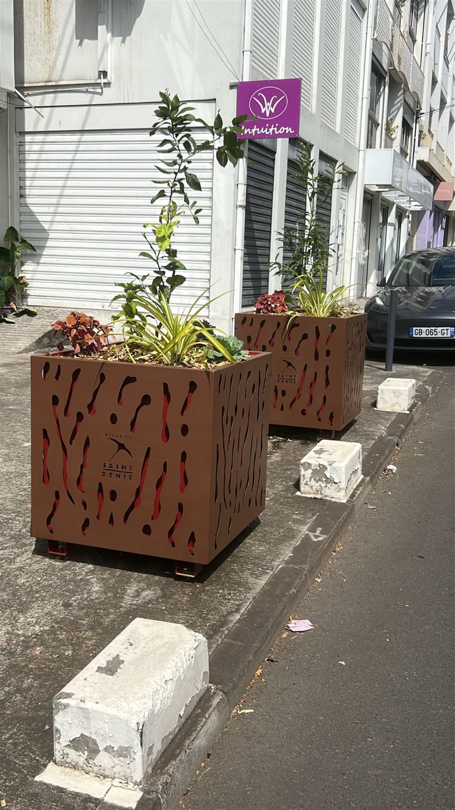 Rue bordée de jardinières couleur rouille et d'un panneau violet à côté d'un bâtiment blanc.