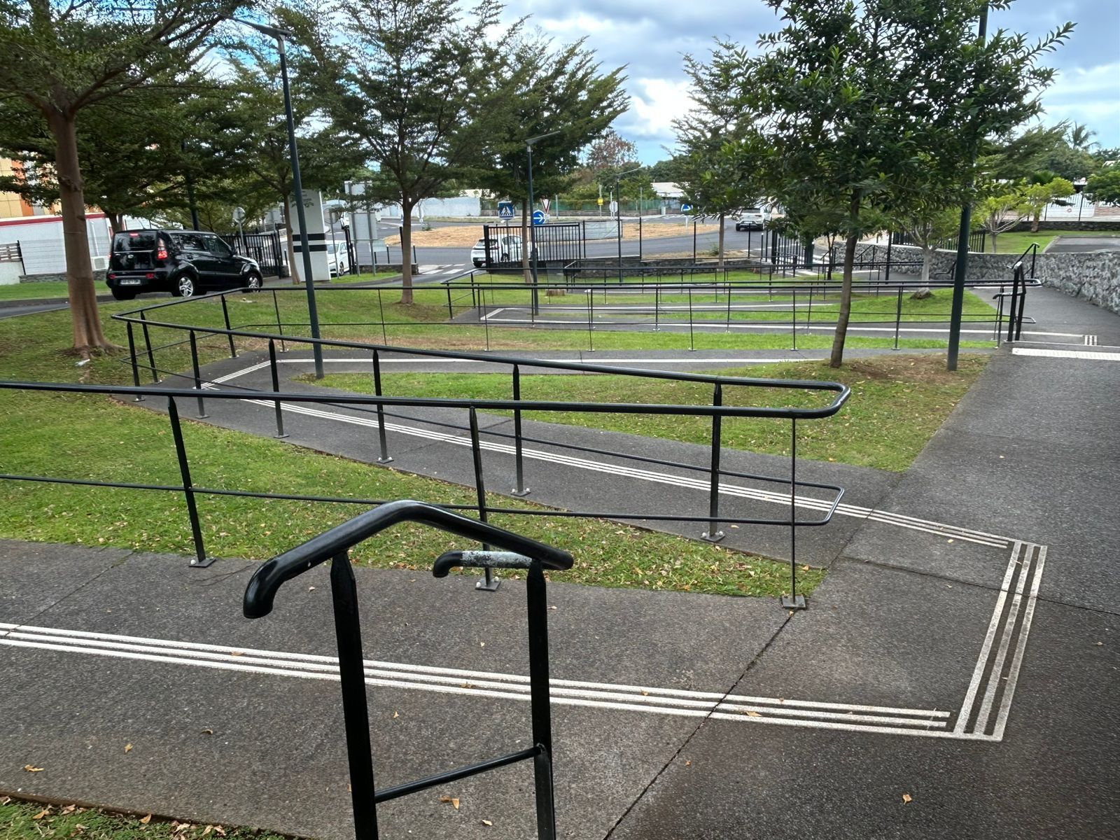 Rampe d'accès pour fauteuil roulant avec barrières noires dans un parc, à côté d'un chemin pavé et d'une zone herbeuse.
