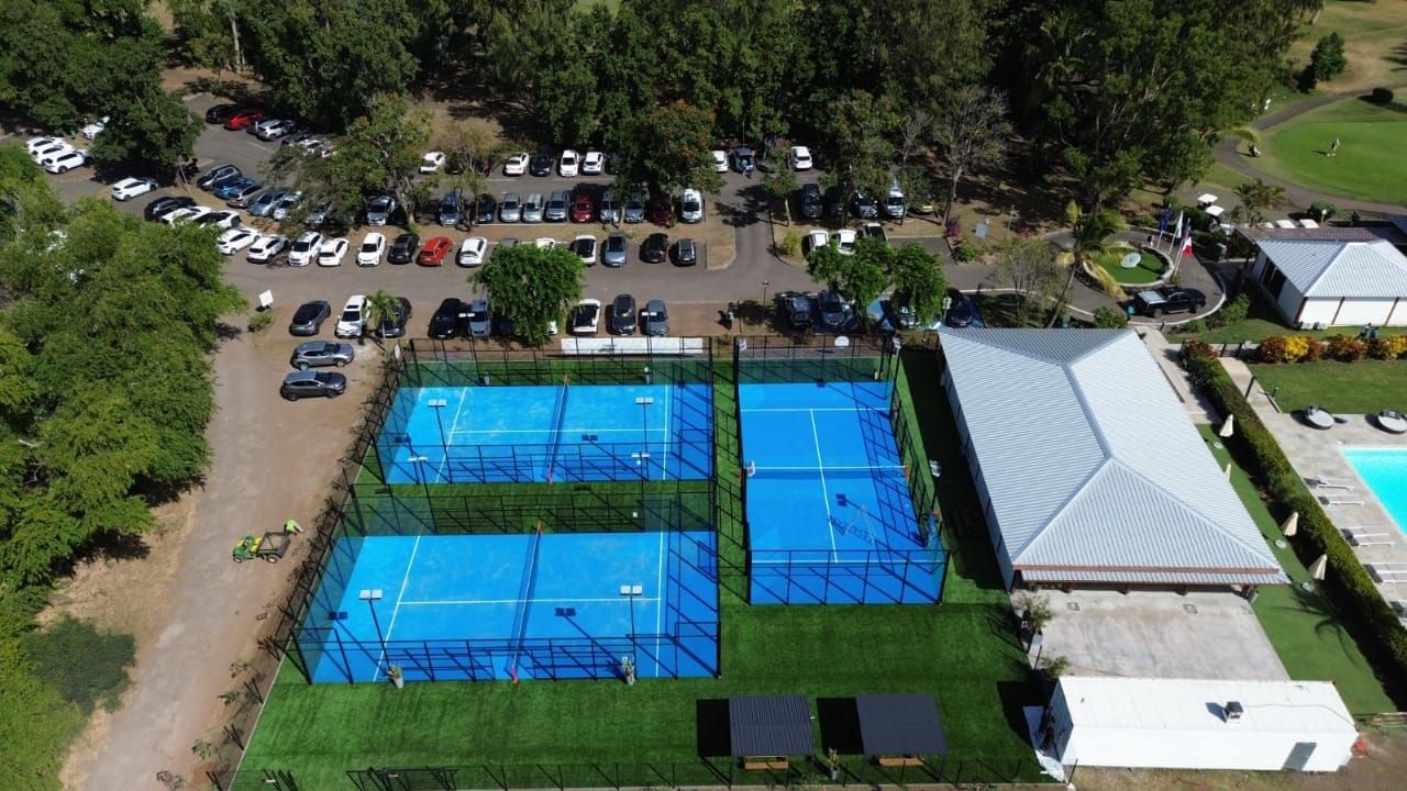 Vue aérienne de quatre courts de padel bleus à côté de voitures garées et de tentes blanches dans un parc bordé d'arbres