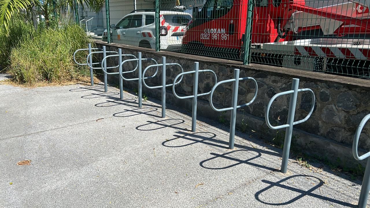 Des supports à vélos en métal longent un chemin de gravier, à côté d'une clôture rouge et de véhicules stationnés.