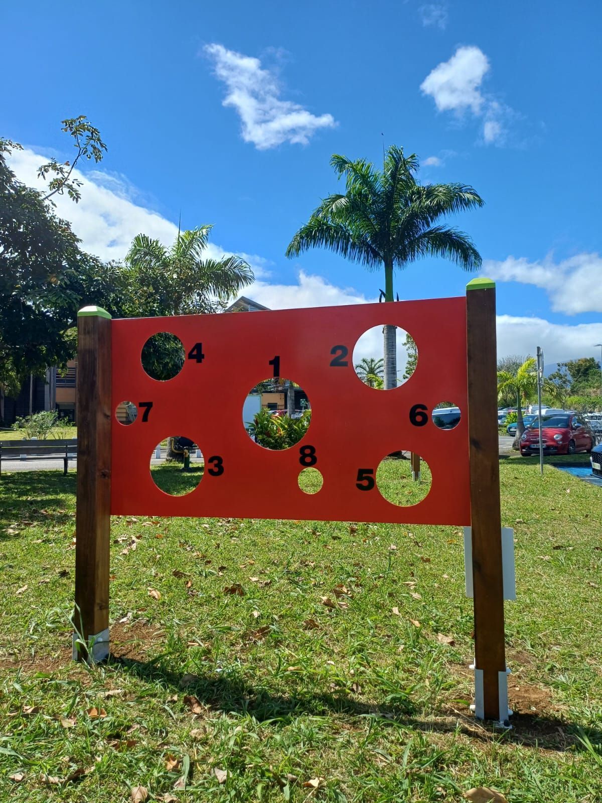 Panneau de jeu rouge avec des trous circulaires numérotés sur un terrain herbeux sous un ciel bleu.