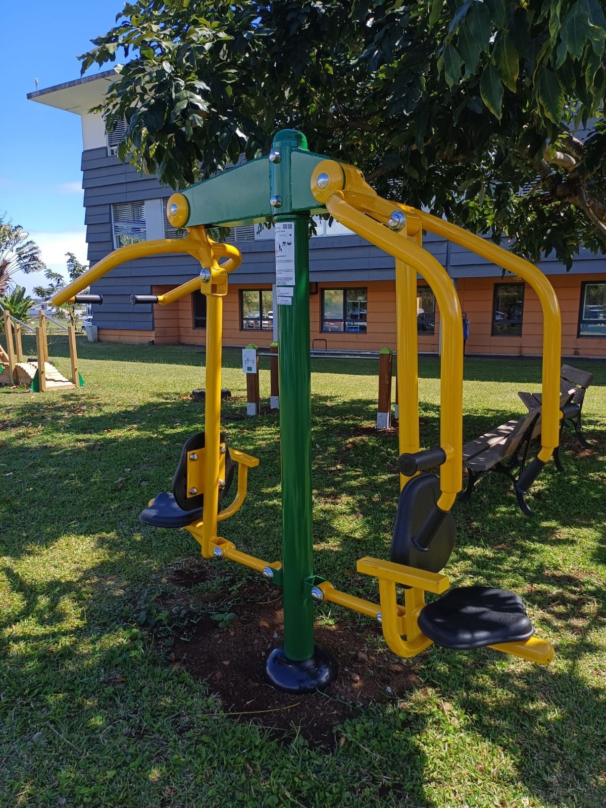 Appareil de fitness jaune en extérieur, dans un parc, à côté d'une pelouse et d'une maison, sous un arbre feuillu.