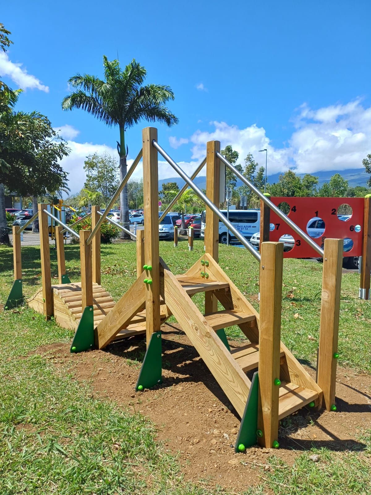 Jeux en bois avec escaliers et rambardes, dans un parc herbeux sous un ciel bleu.