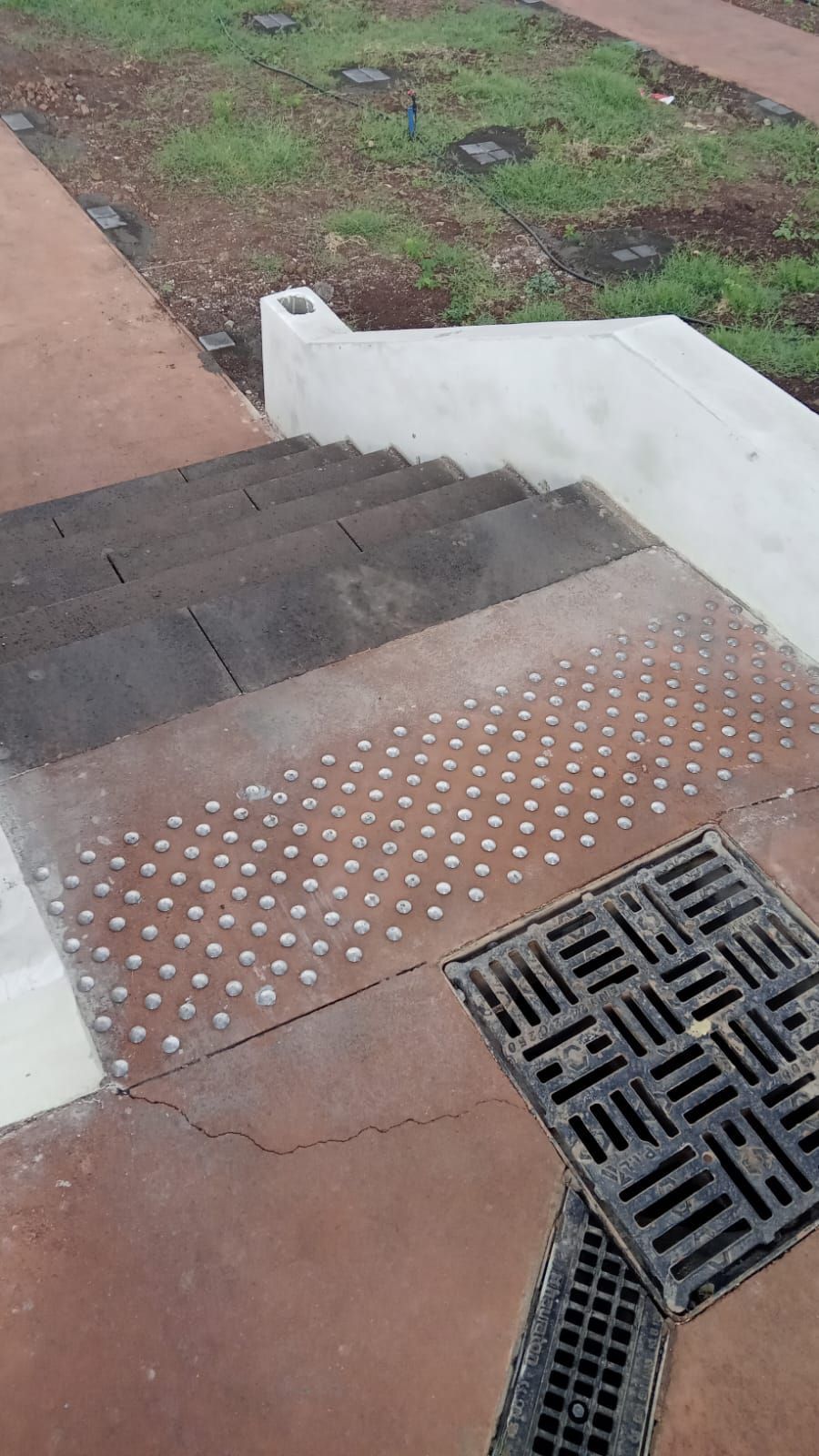 Escalier extérieur avec revêtement tactile, marches foncées et grille d'évacuation des eaux pluviales à côté d'une rampe en béton.