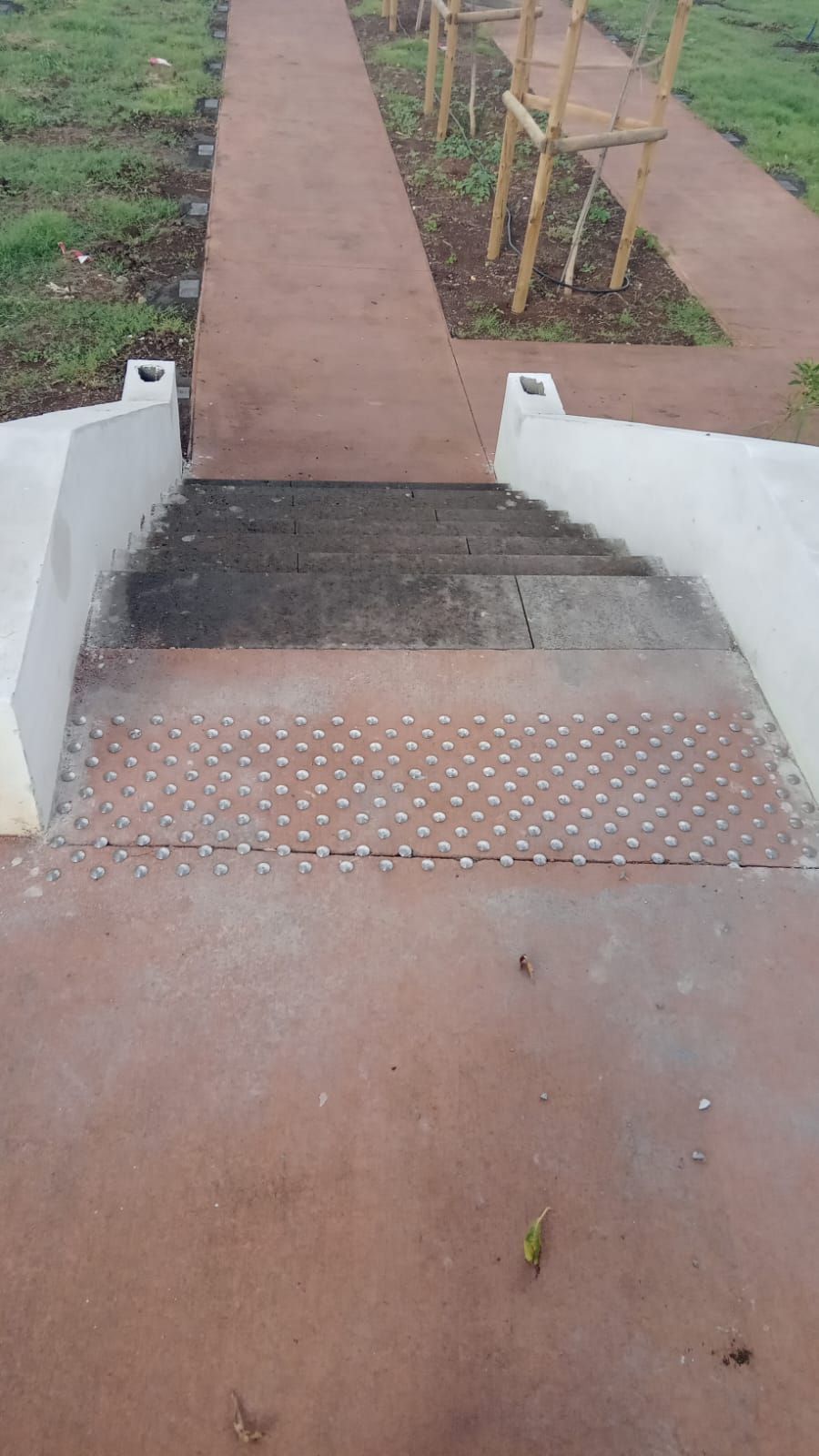 Petit escalier avec dalles tactiles et murs latéraux blancs menant à un chemin marron dans un parc.