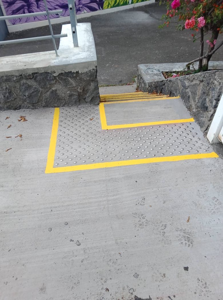 Marche et rampe d'accès balisées en jaune à côté d'un chemin en béton et d'un mur en pierre.