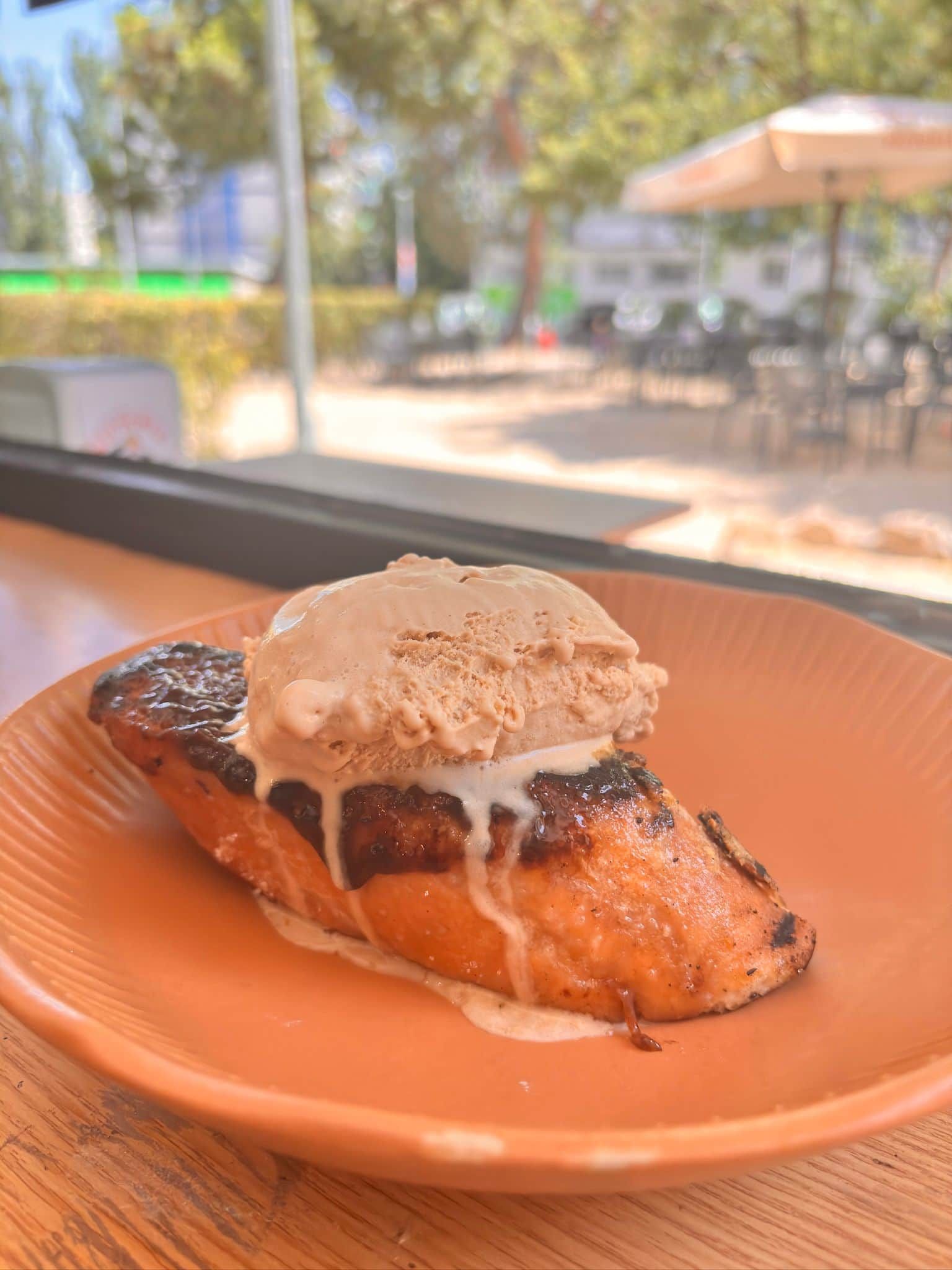 Postre a la parrilla coronado con una bola de helado en un plato naranja, colocado junto a una ventana con vistas a un patio.