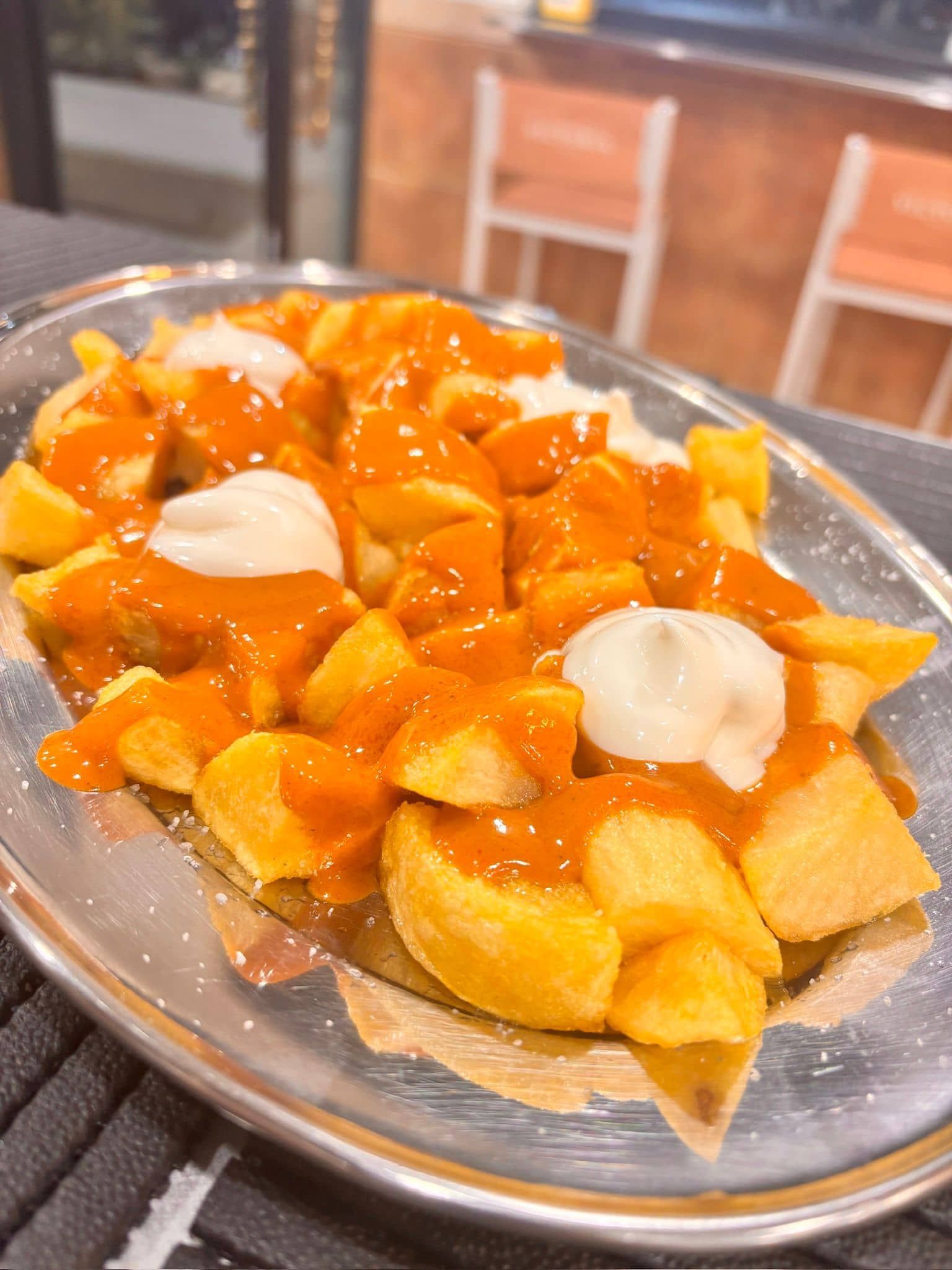 Patatas bravas: cubos de patata fritos cubiertos con salsa roja y alioli blanco, servidos en una bandeja de plata.