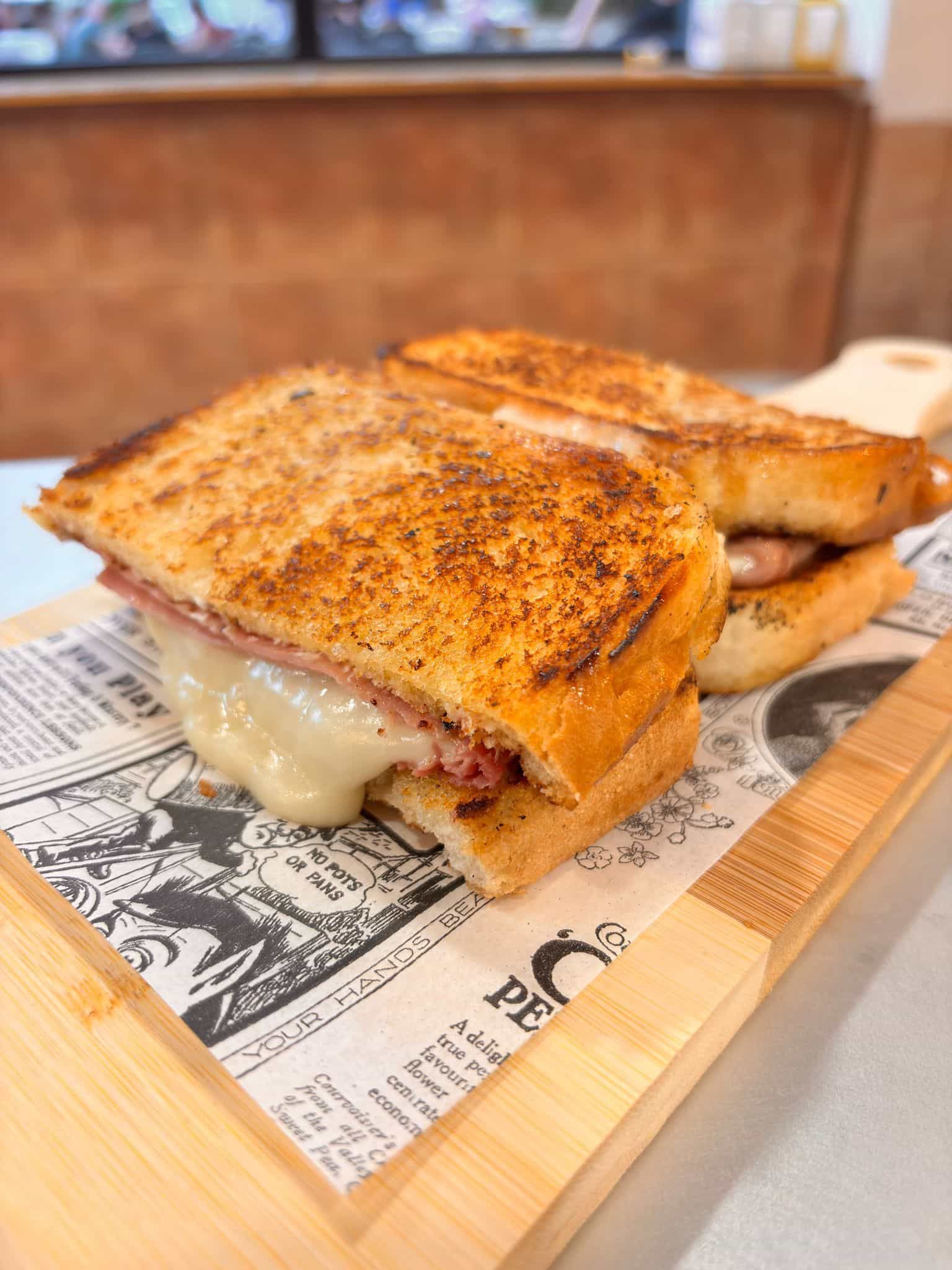 Sándwich a la plancha con queso fundido sobre una tabla de madera, apoyado sobre papel de periódico.