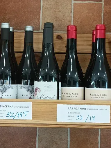 Botellas de vino tinto en un estante de madera; etiquetas de precio debajo.