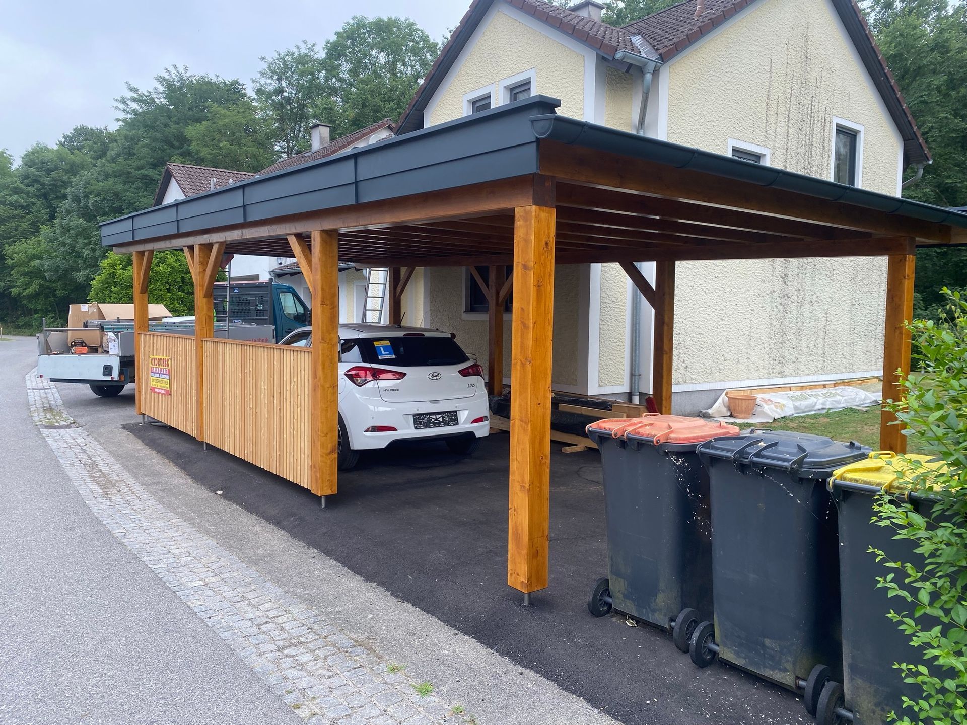 Holzcarport mit einem darin geparkten weißen Auto. Neben der Einfahrt stehen schwarze Mülltonnen.