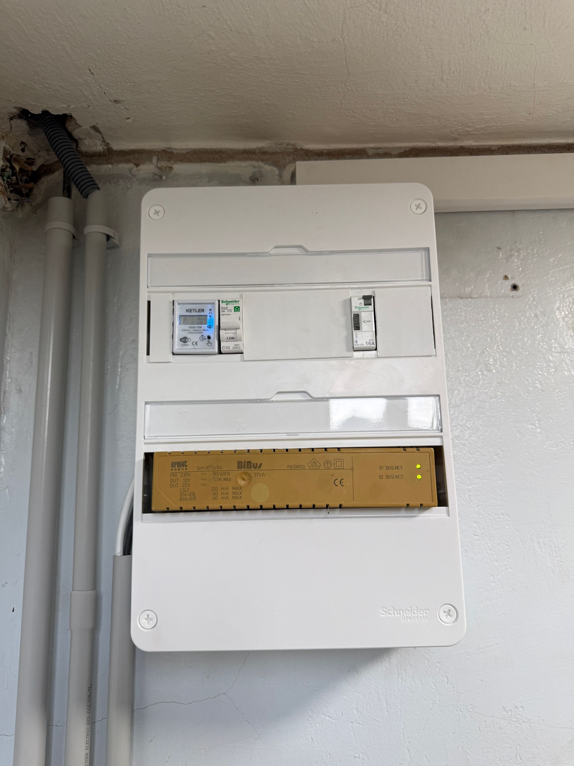 Boîtier de compteur électrique blanc fixé sur un mur gris, dans un garage.