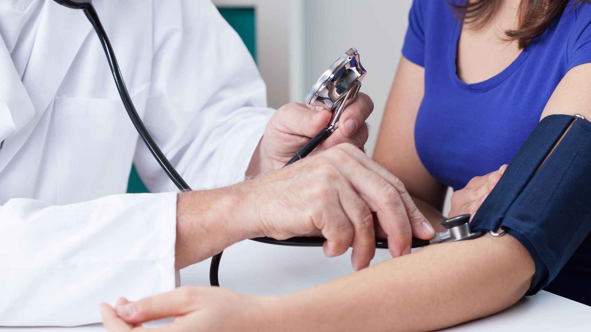 Médico aferindo a pressão arterial de um paciente com estetoscópio e esfigmomanômetro em um consultório.