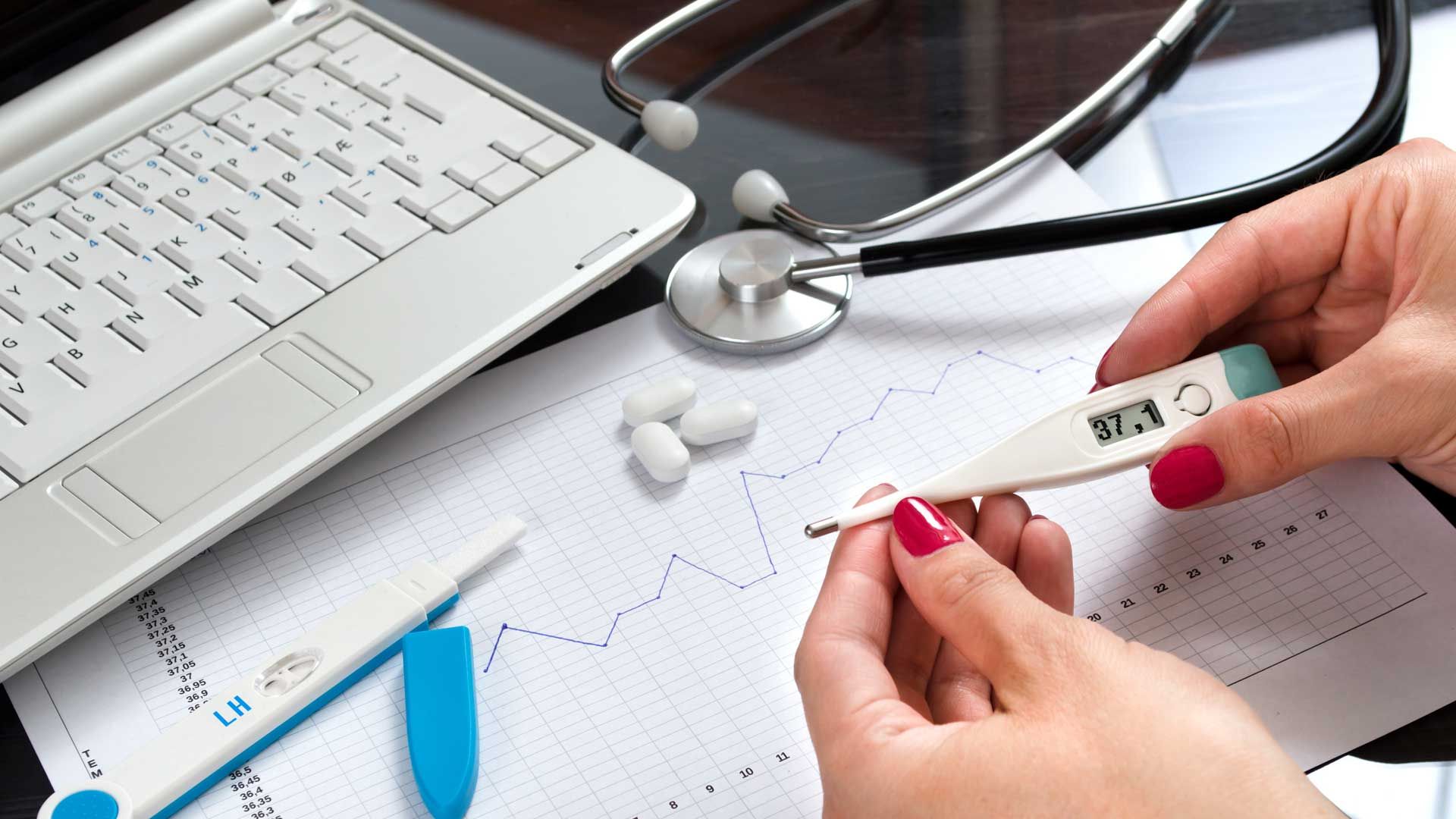 Mãos segurando um termômetro, ao lado de um laptop, estetoscópio, comprimidos e prontuário médico.