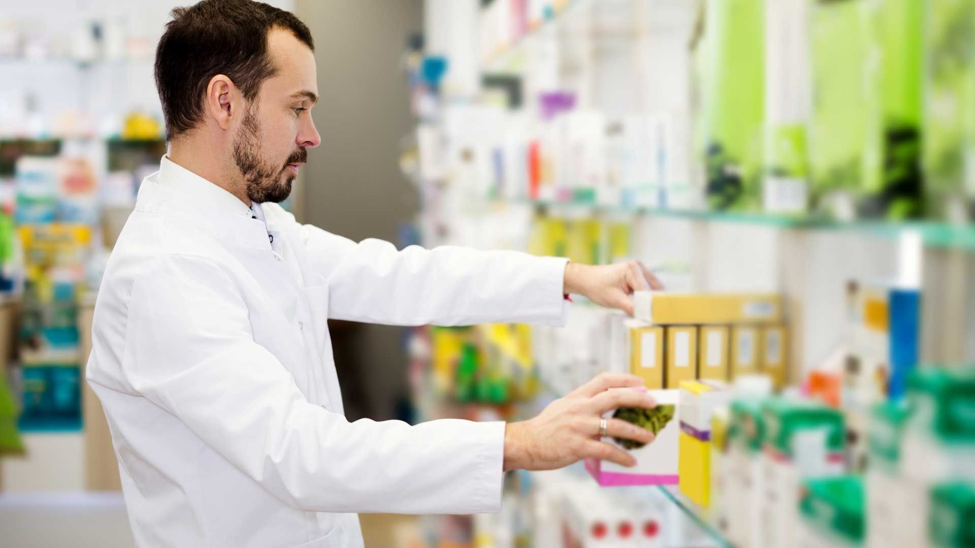 Farmacêutico de jaleco branco organizando medicamentos em uma prateleira da farmácia.