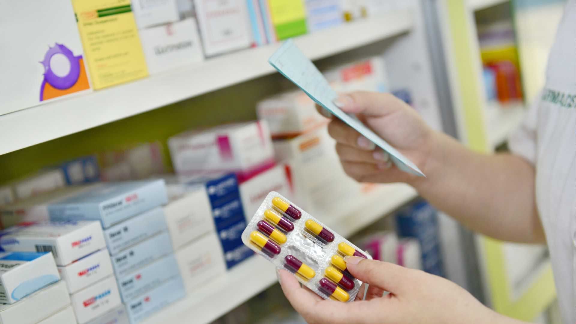Farmacêutico de jaleco branco lendo uma receita, segurando uma cartela de comprimidos em frente às prateleiras.