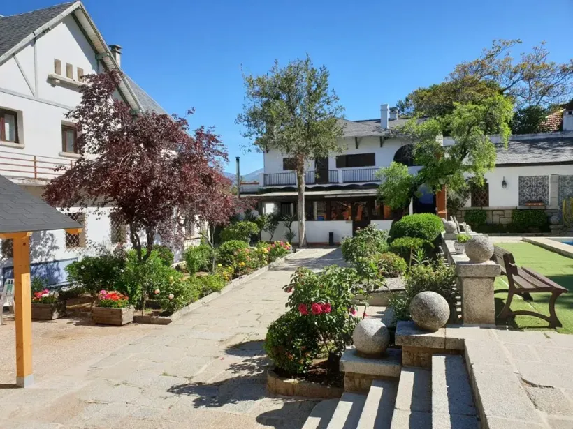 Un patio con un sendero de piedra bordeado de flores conduce a edificios blancos bajo un cielo azul. Se ven un árbol de hojas rojas y arbustos bien cuidados.