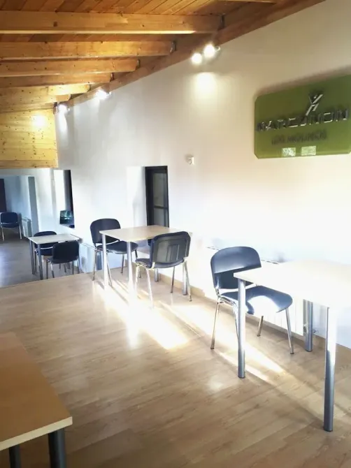 Interior de una sala luminosa con mesas y sillas. Se aprecia el techo de madera, las paredes blancas y un rótulo comercial verde.