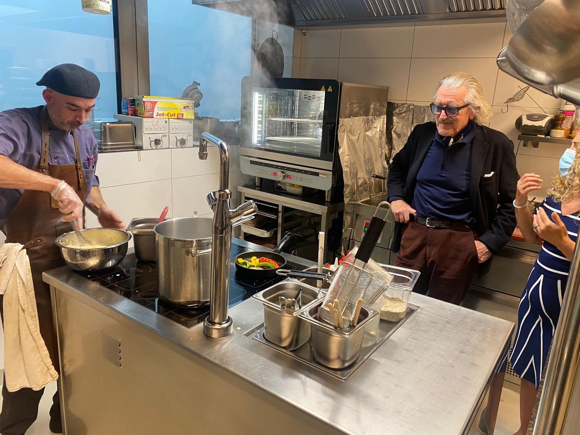 Zwei Männer kochen in einer Küche Essen, während eine Frau zusieht.