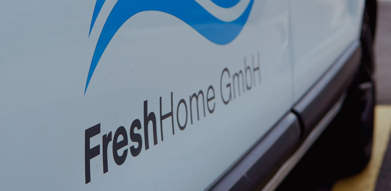Seitenansicht eines weißen Fahrzeugs mit einem blauen Logo und dem schwarzen Schriftzug „FreshHome GmbH“ auf der Tür.