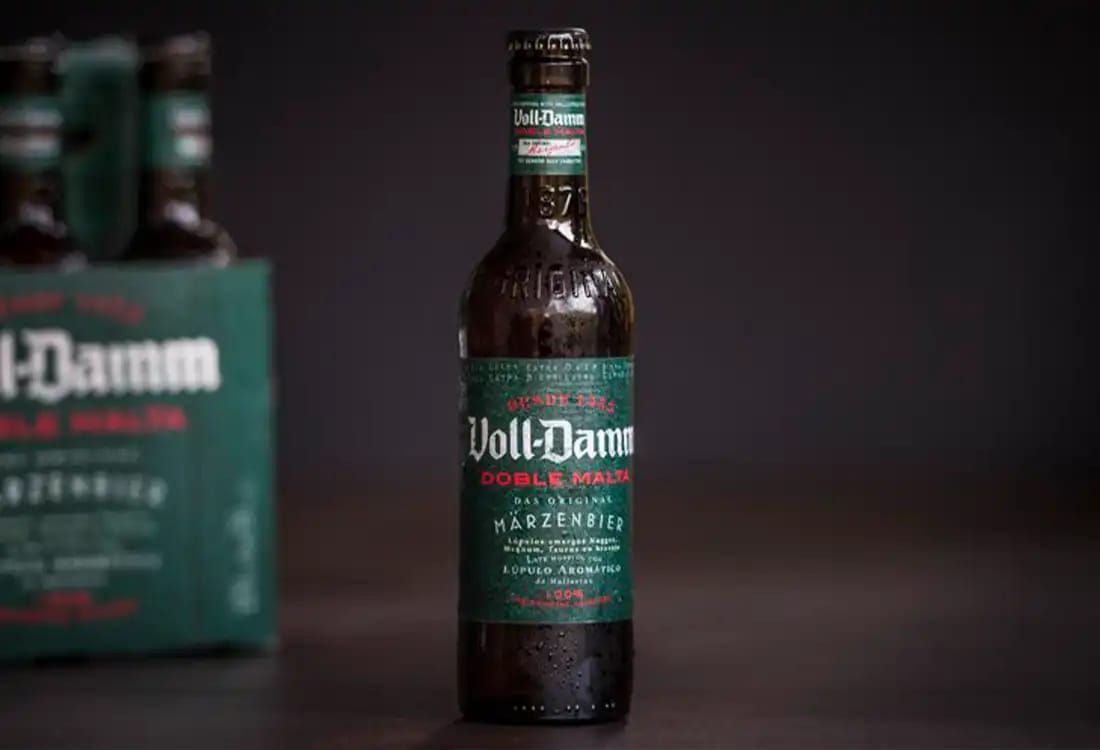 Botella individual de cerveza Voll-Damm Doble Malta con etiqueta, junto a un pack de seis, fondo oscuro.