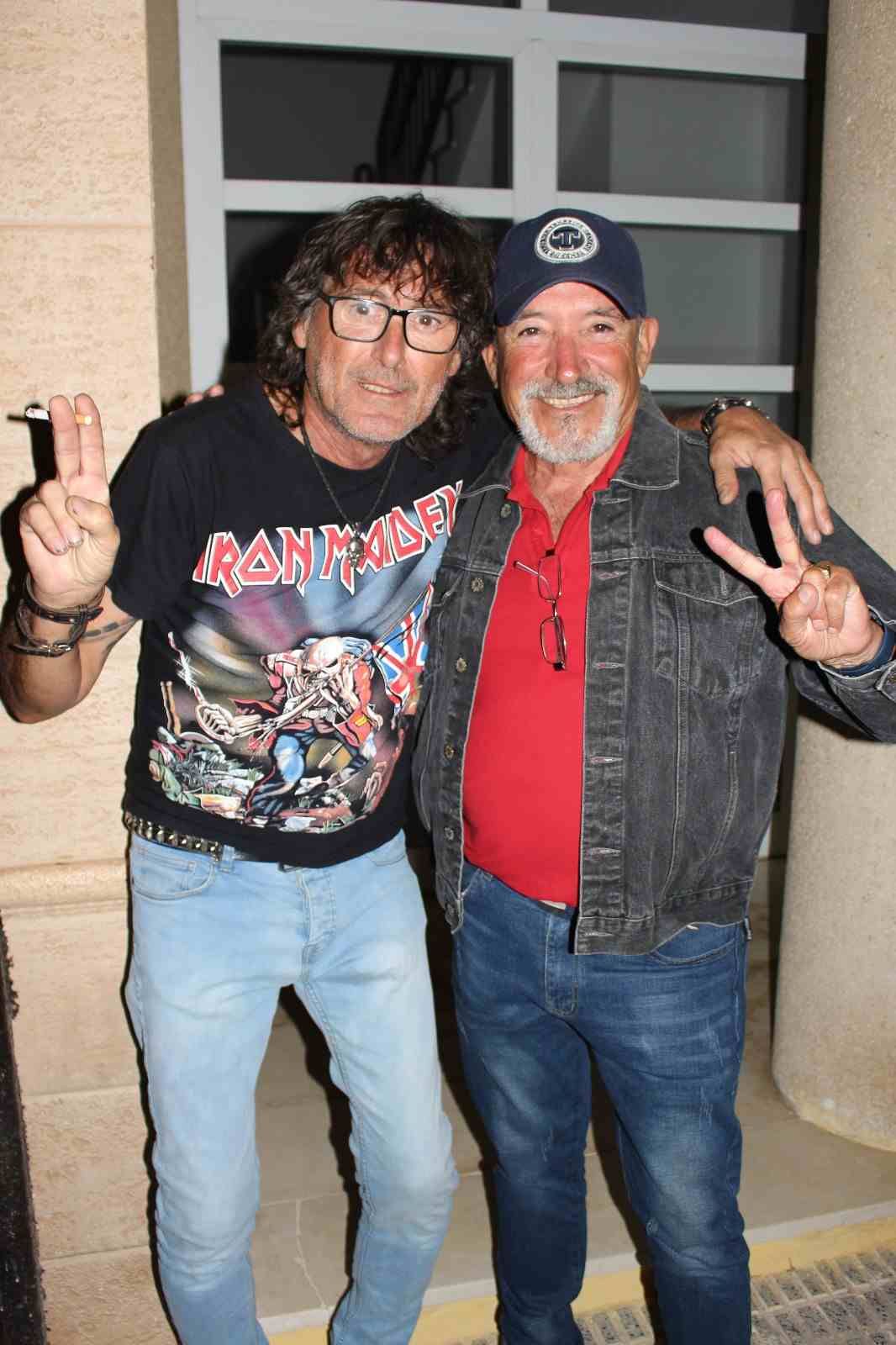 Dos hombres sonriendo, posando. Uno lleva una camiseta de Iron Maiden, gafas y el símbolo de la paz. El otro lleva una chaqueta vaquera, sombrero, camiseta roja y el símbolo de la paz.