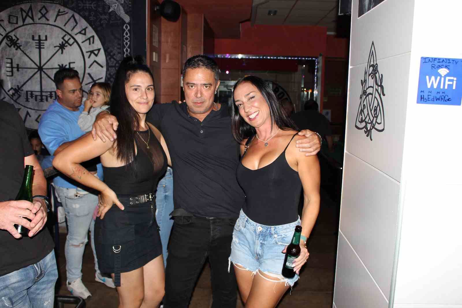 Un hombre abraza a dos mujeres en un bar. Sonríen, sosteniendo bebidas. Interior con clientes.