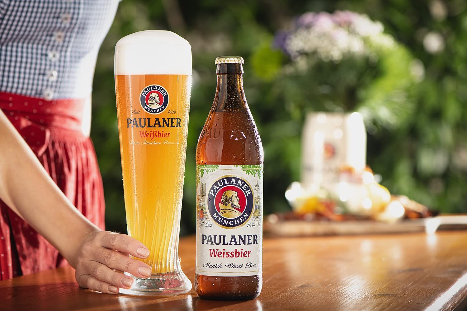 Un vaso alto de cerveza Paulaner junto a una botella sobre una mesa con la mano de una persona cerca.