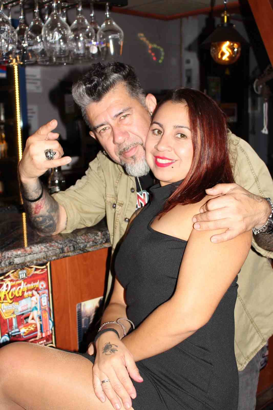 Un hombre con tatuajes abraza a una mujer en un bar; hace un gesto con la mano. Ambos sonríen.