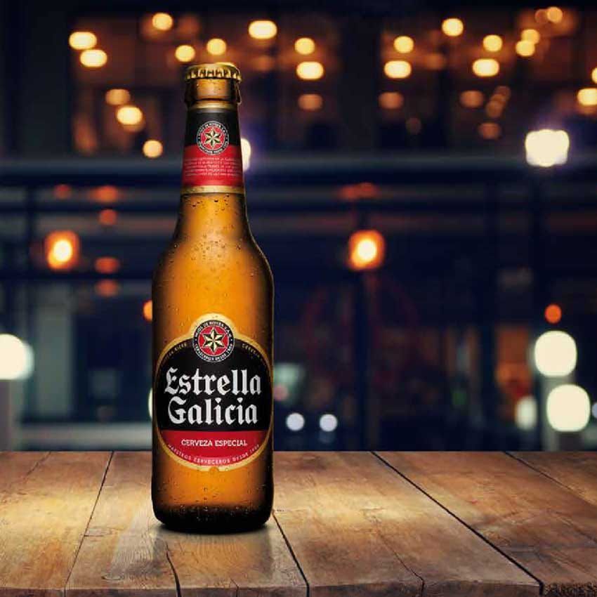 Botella de cerveza Estrella Galicia sobre una mesa de madera, sobre un fondo borroso de luces.