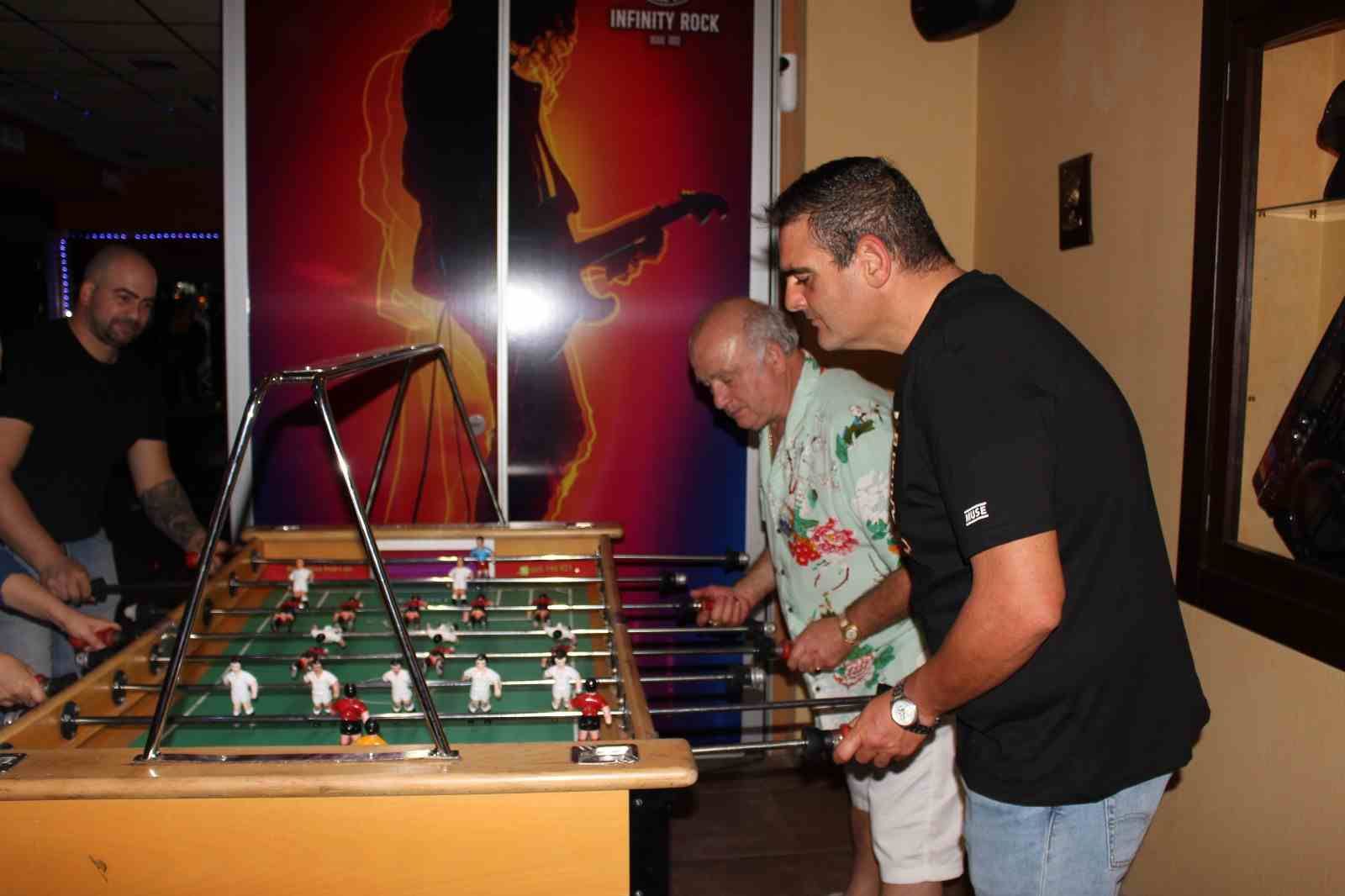 Gente jugando al futbolín en un bar. Dos hombres concentrados en el partido, otros observando. Un cartel colorido al fondo.