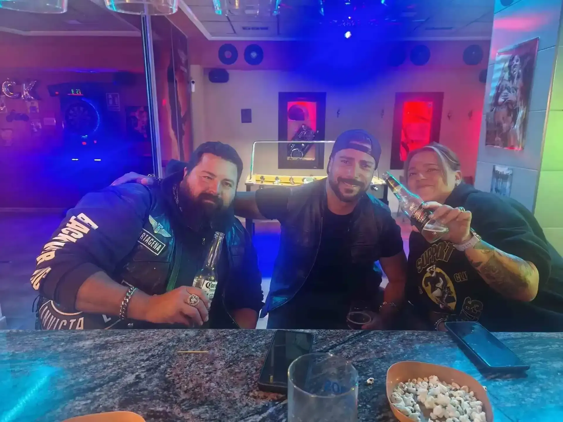 Tres personas en un bar, alzando botellas de cerveza. Interior oscuro e iluminado con luz azul.